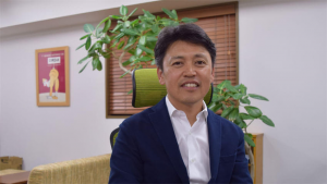 Portrait of a man in a navy blazer and white shirt, seated in a modern office with greenery behind him.